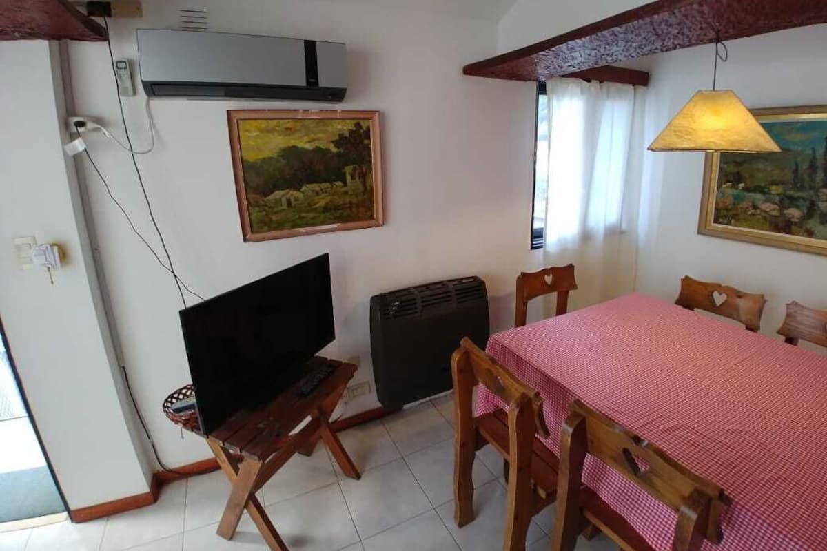 Uma sala de jantar com toalha xadrez vermelha, cadeiras de madeira, uma televisão sobre um móvel e quadros nas paredes.