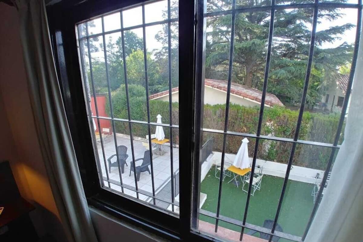 Uma janela com grades, vista para um pátio externo com mesas e cadeiras, e um gramado verde.