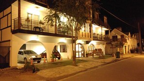 A two-story building with a balcony on the upper floor, a car parked in the driveway, and a small garden area with potted plants.