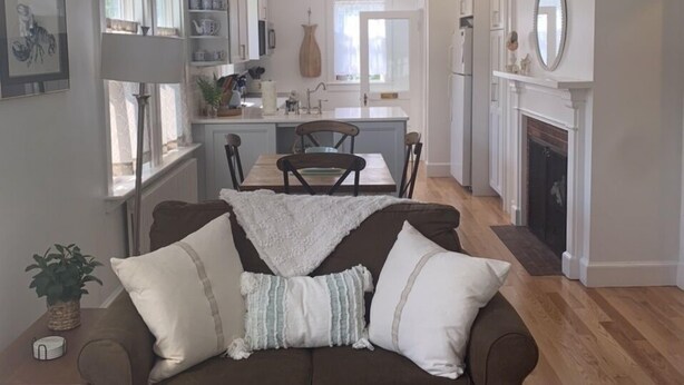 Uma sala de estar com um sofá marrom, um espaço de jantar com mesa e cadeiras, uma cozinha com armários brancos e uma lareira.