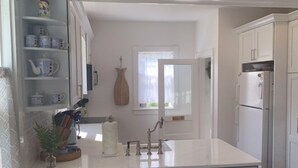 Cozinha com bancada branca, pia e geladeira. Há uma janela com vista para o exterior.