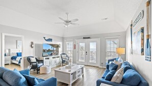 Ein geräumiges Wohnzimmer mit einer großen Couch, einem Couchtisch und einem Fernseher. Eine Tür mit der Aufschrift „Welcome“ und ein Schild mit der Aufschrift „Beach House“.