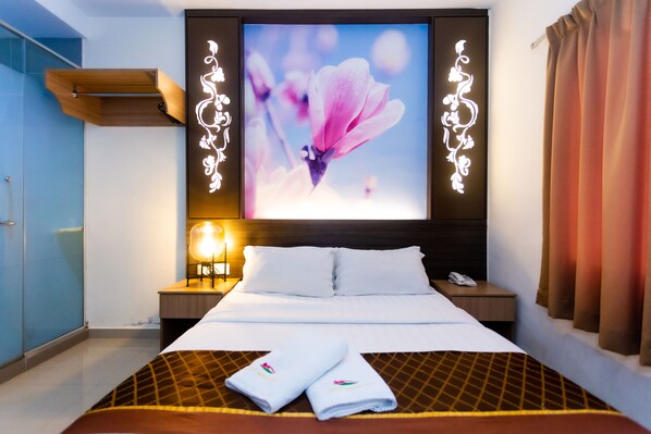 Una habitación de hotel con una cama grande, una mesita de noche con lámpara y un televisor de pared que muestra una imagen floral.