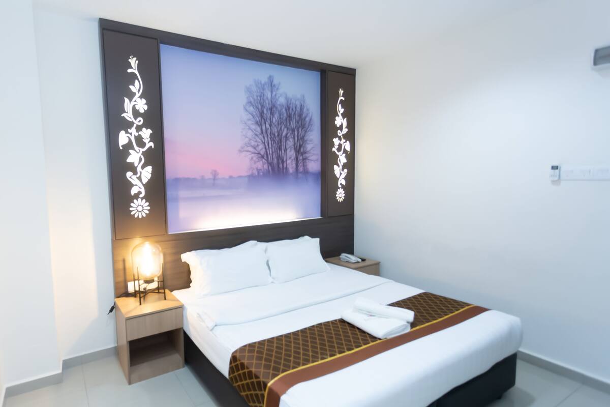 A hotel room with a bed, bedside table, lamp, and a large picture on the wall.