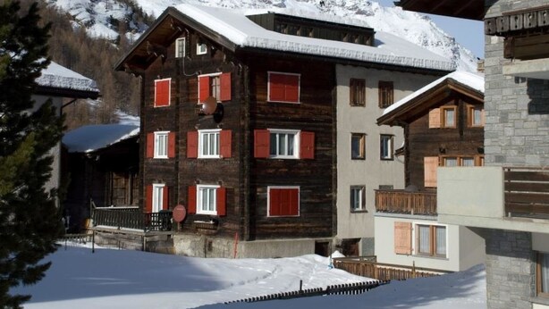 Un immeuble à plusieurs étages, doté d’une façade en bois et de volets rouges, entouré de montagnes enneigées.