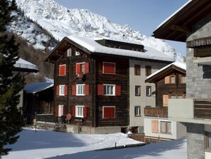 Un edificio de varios pisos con fachada de madera y contraventanas rojas, rodeado de montañas nevadas.