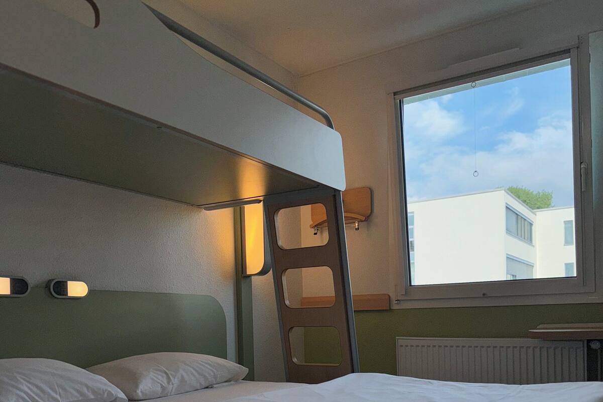 Un lit simple avec une literie blanche, dans une pièce dotée d’une fenêtre, d’un radiateur et d’un luminaire fixé au mur.
