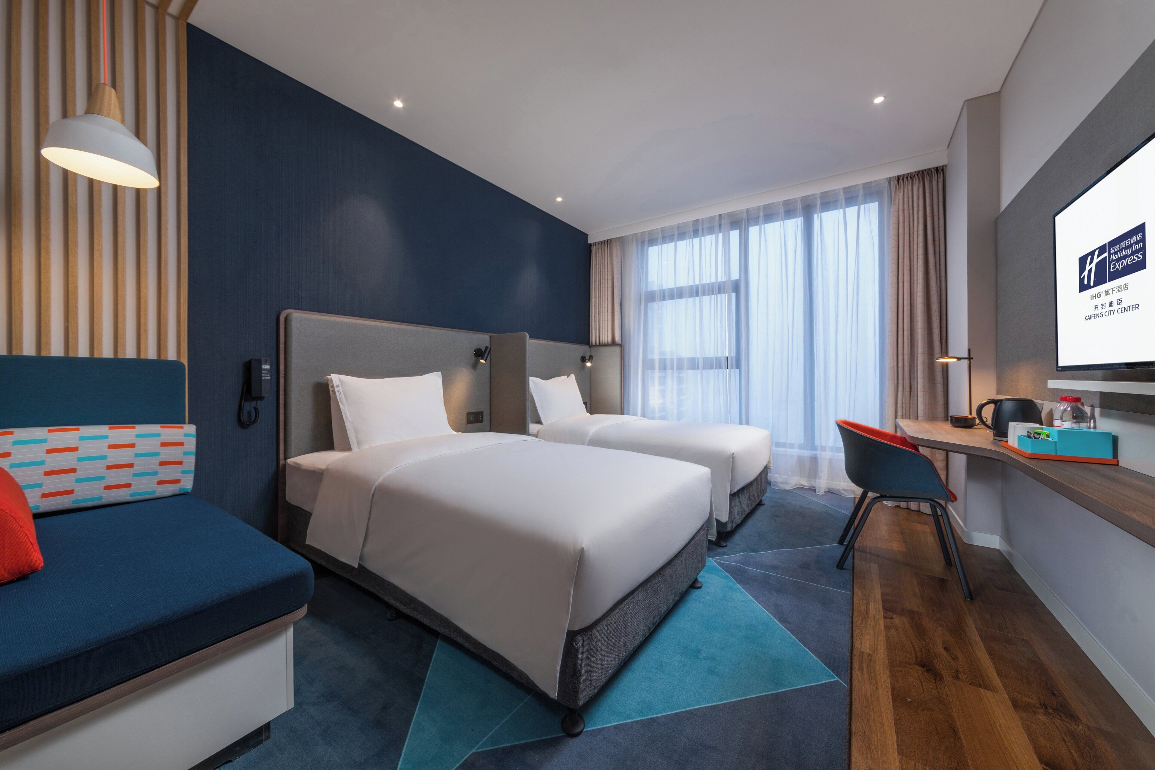 Habitación de hotel con dos camas, un escritorio, una silla y una pantalla de televisión que muestra el nombre del hotel.