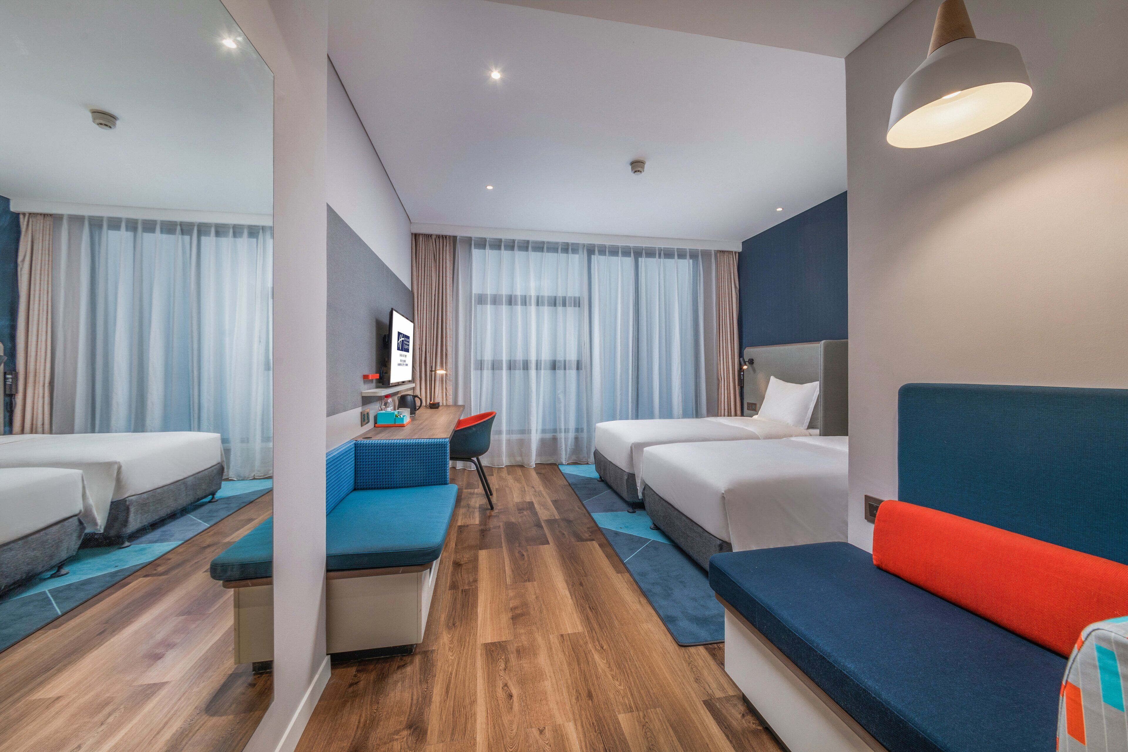 A modern hotel room with a wooden floor, a sofa, two beds, a desk with a chair, and a TV.