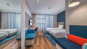 A modern hotel room with a wooden floor, a sofa, two beds, a desk with a chair, and a TV.