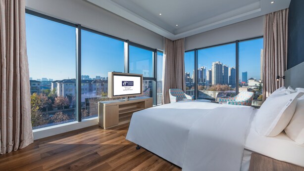 A modern hotel room with a large bed, a desk, and a TV. The room has large windows offering a city view.