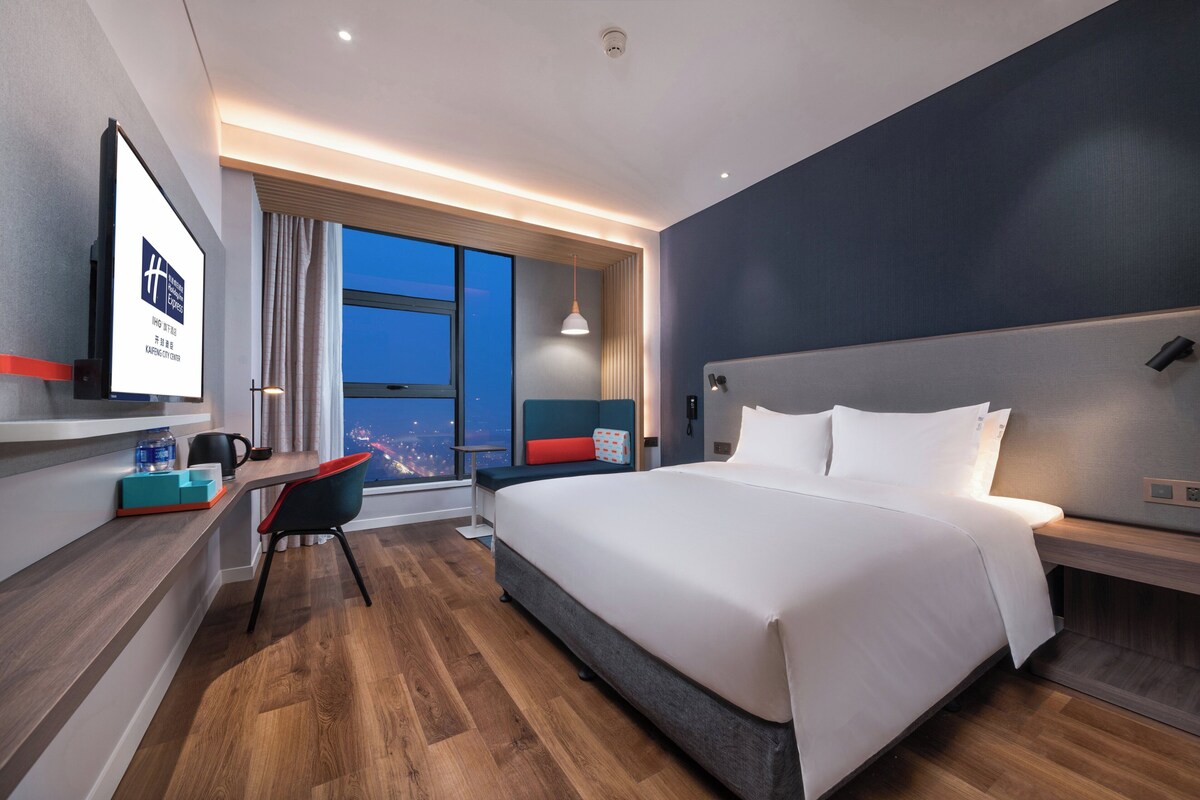 A modern hotel room with a large bed, a desk with a chair, a large window, and a city view.