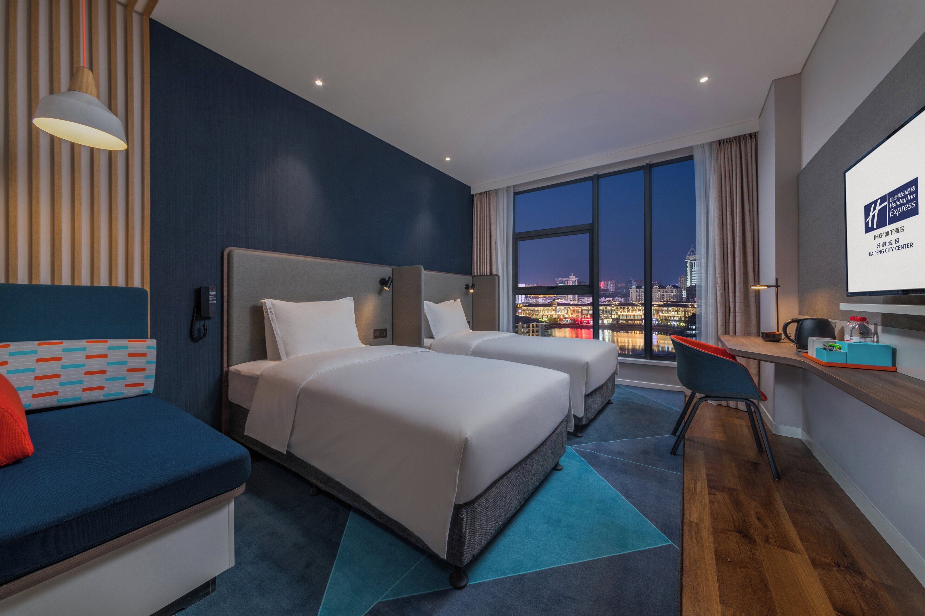 Une chambre d’hôtel avec deux lits, un bureau, une vue sur la ville la nuit et une grande fenêtre.