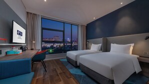 A hotel room with two beds, a desk, a chair, and a large window offering a city view.