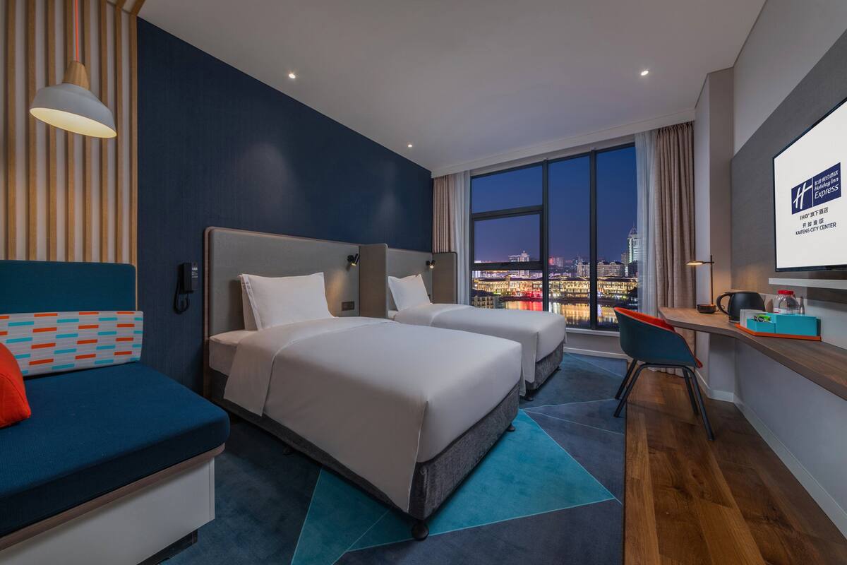 A hotel room with two beds, a desk, a chair, and a large window offering a city view.