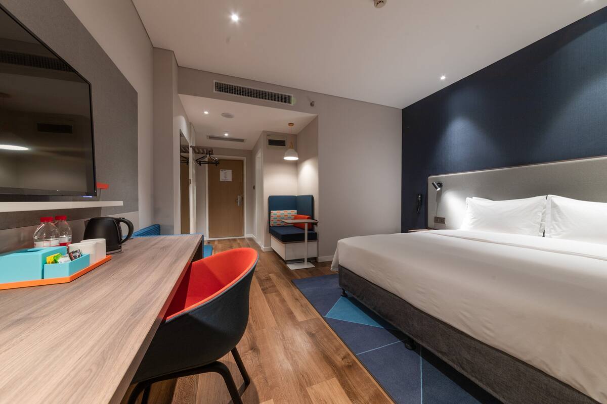 A hotel room with a bed, a desk with a chair, a TV, and a kitchenette area.
