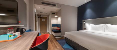 A hotel room with a bed, a desk with a chair, a TV, and a kitchenette area.