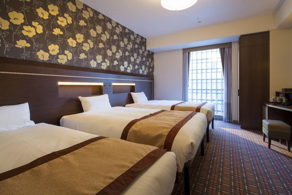 Una habitación de hotel con tres camas, un ventanal grande con cortinas y una alfombra estampada.