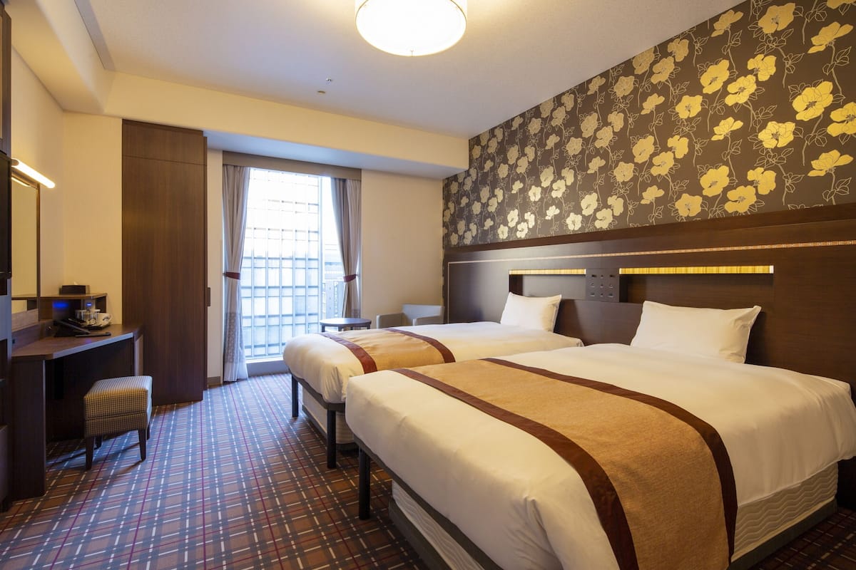 Habitación de hotel con dos camas, un escritorio, una silla y un ventanal con cortinas.