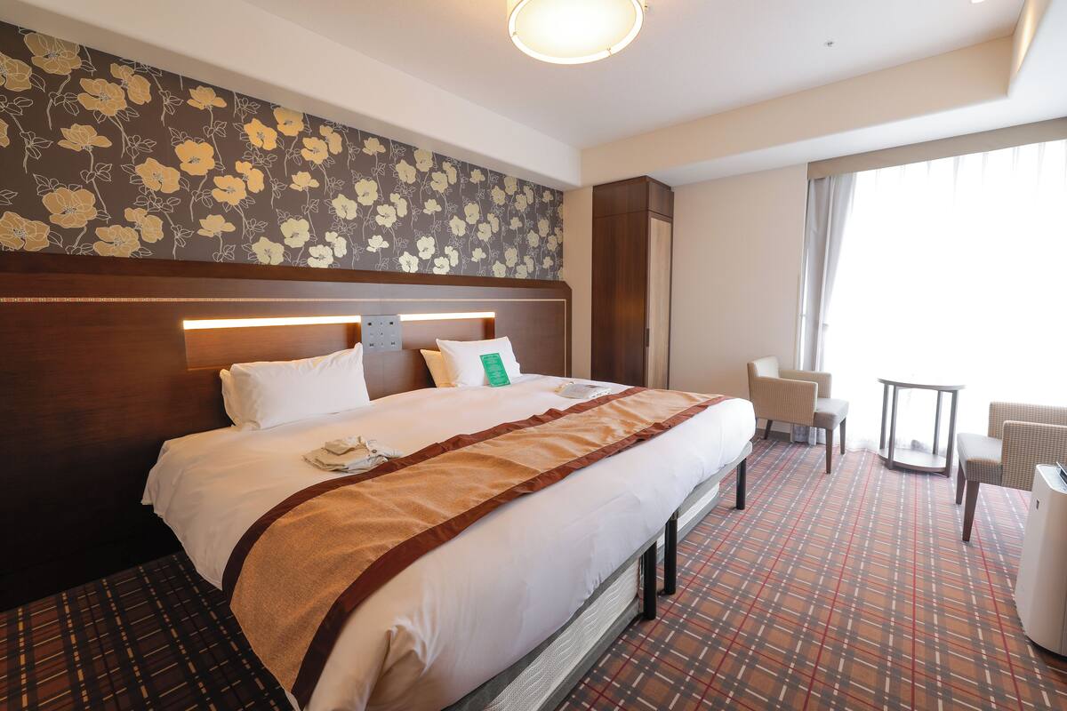Habitación de hotel con una cama grande, cabecera con lámpara, papel tapiz floral, una mesita y una silla.