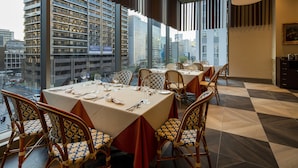 A dining area with tables set for four, wicker chairs, and a city view through large windows.