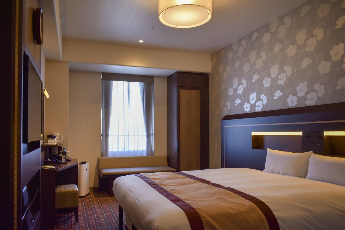 Habitación de hotel con una cama grande, un escritorio con silla, un pequeño espacio de descanso y una ventana con cortinas.