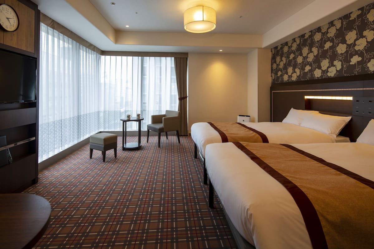 Habitación de hotel con dos camas, televisión, una mesita, una silla y vistas a la ciudad a través de amplias ventanas.