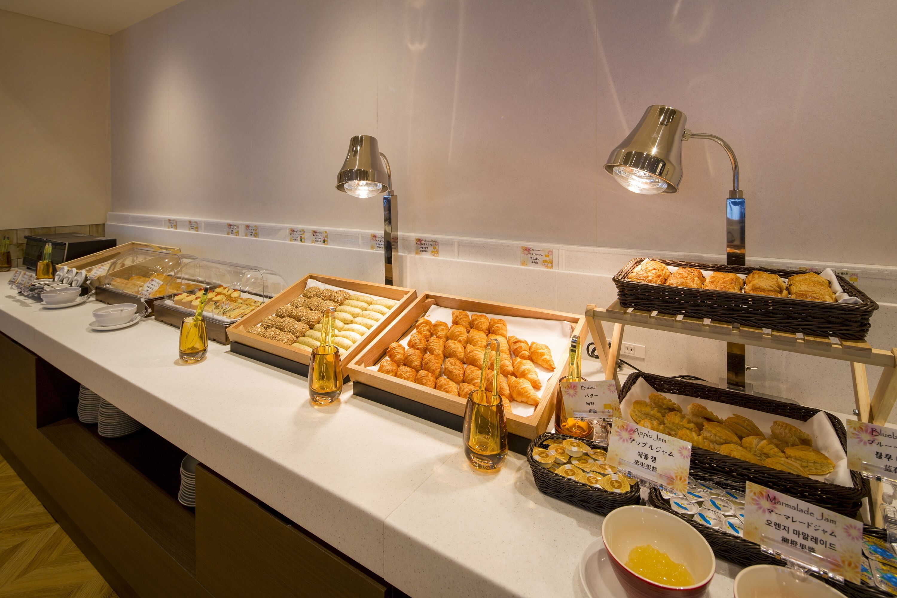 Un bufé con diversos alimentos y acompañamientos, como pasteles, sándwiches y mermeladas, con etiquetas y una lámpara.