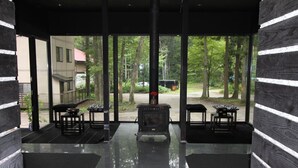 A modern, enclosed outdoor space with a fireplace, seating area, and large windows offering a view of a forested area. - Alpine Lodge (Hakuba)