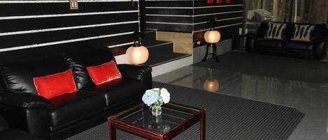 Una sala de estar moderna con un sofá de cuero negro, cojines rojos, una mesa de centro de vidrio con un jarrón de flores y una pared a rayas negras y blancas con detalles rojos.