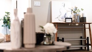 Ein Zimmer mit einem Holztisch, einer weißen Vase mit Blumen, einem gerahmten Bild an der Wand und einem kleinen Regal mit Tassen und einer Vase.