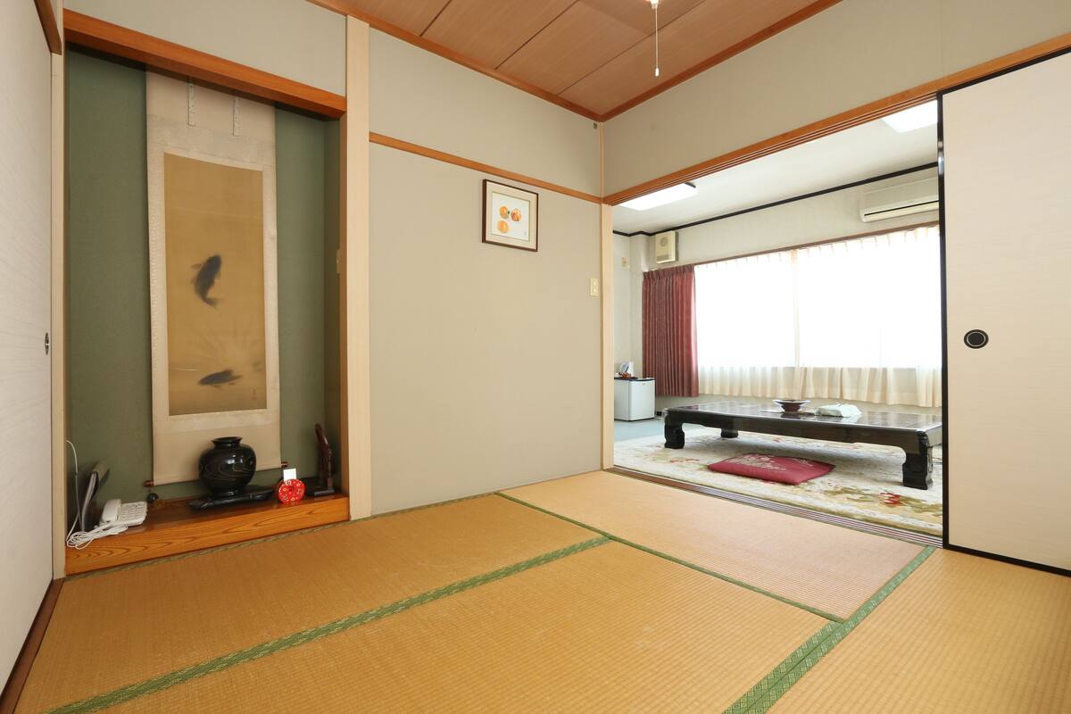 Perinteinen japanilainen huone, jossa on tatamimatto, shoji-liukuovi, futon-patja ja pieni pöytä, jonka päällä on maljakko ja puhelin.