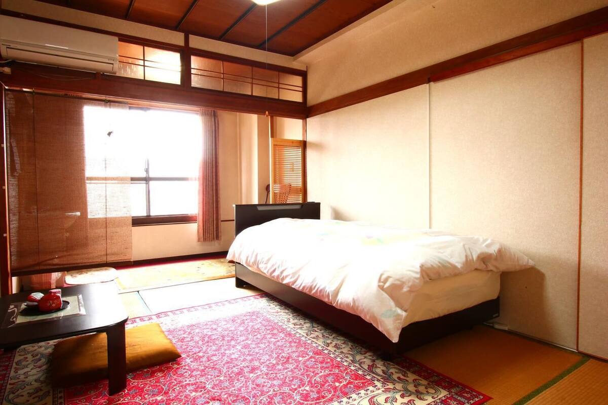 Perinteinen japanilaistyylinen makuuhuone, jossa on tatamimatto, matala sänky, pieni pöytä, jonka päällä on punainen alusta, ja liukuovi, josta on näkymä ulos.