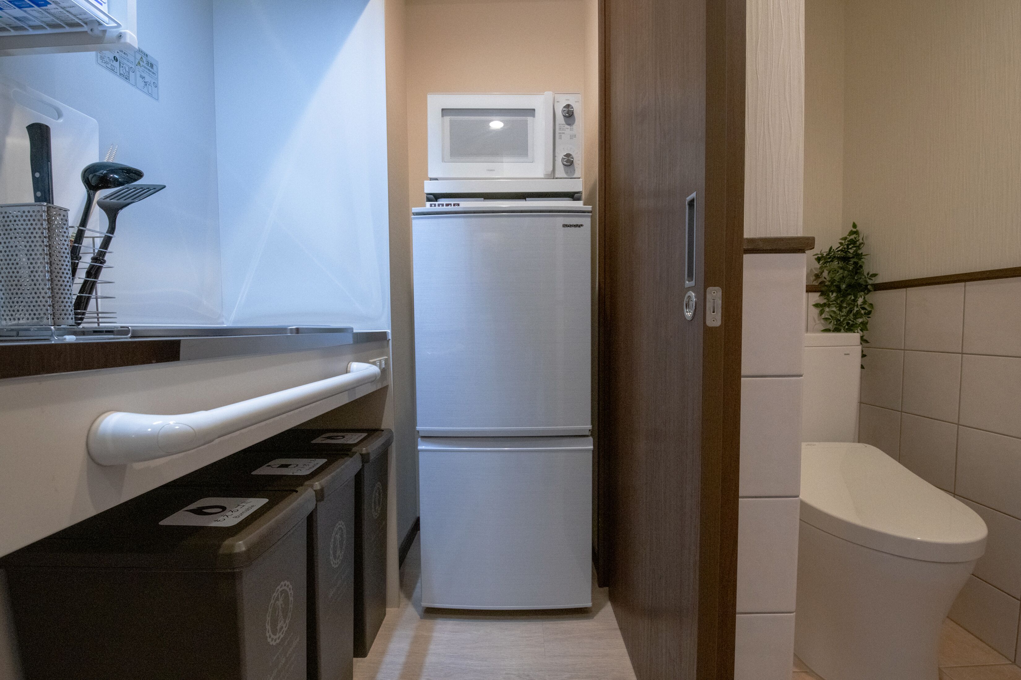 Una cocina compacta con un refrigerador blanco, un microondas y un lavamanos con grifo. Hay armarios debajo de la encimera y una pequeña planta en la pared.