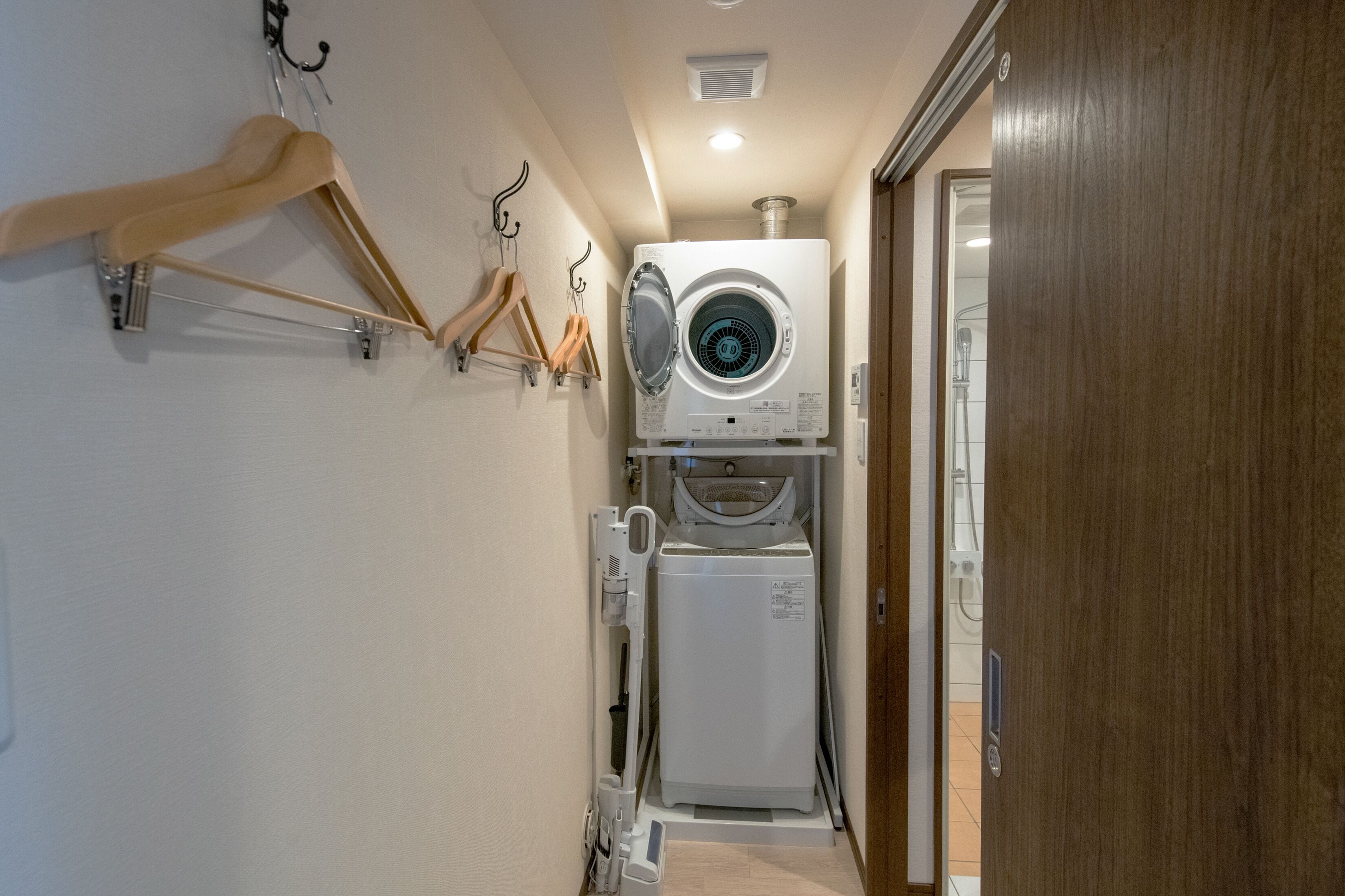 A compact laundry room with a stacked washer and dryer, wooden hangers on a rack, and a wooden door.