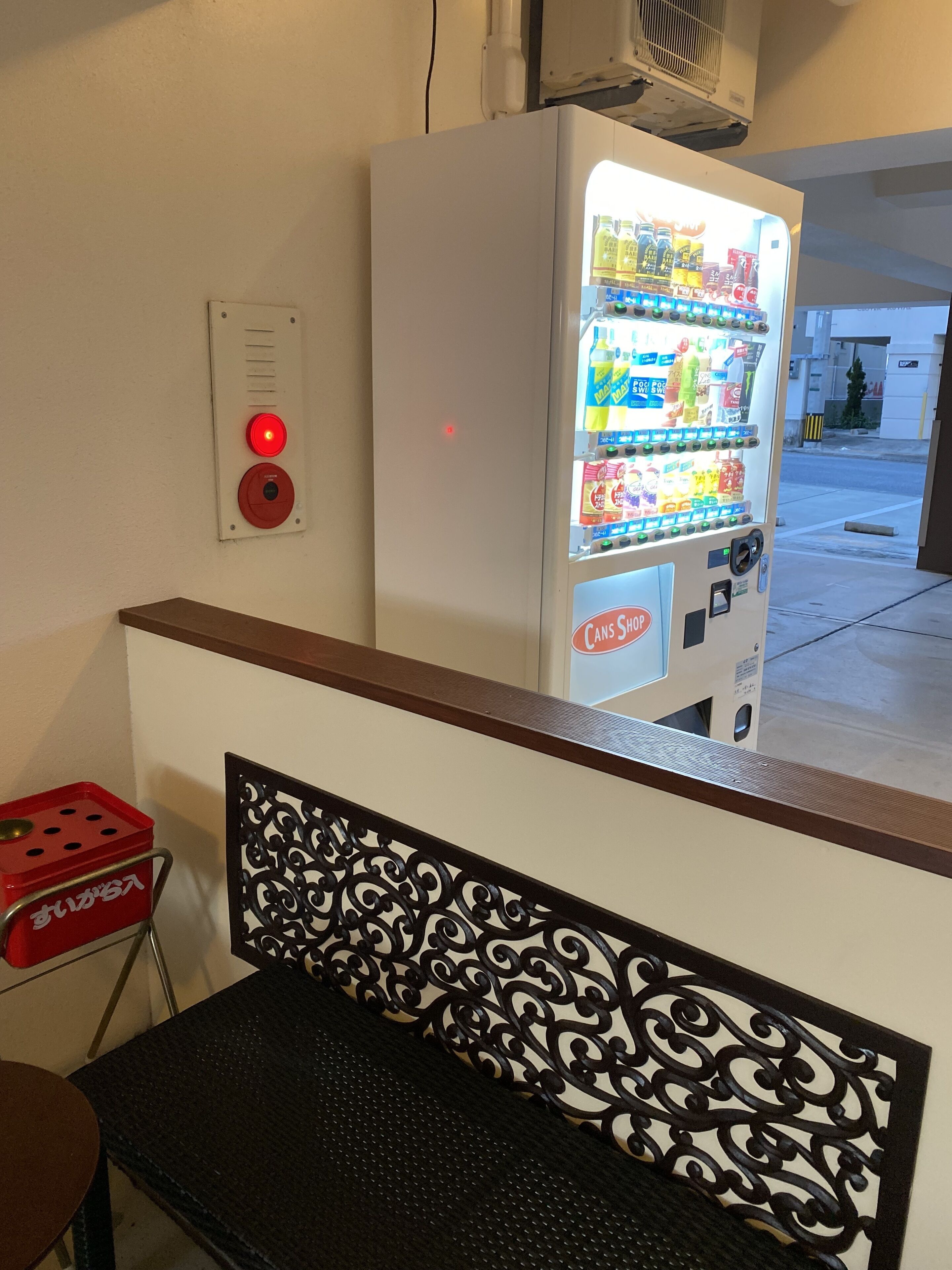 A vending machine with a variety of beverages and snacks, a red emergency button, and a metal chair with a decorative backrest.