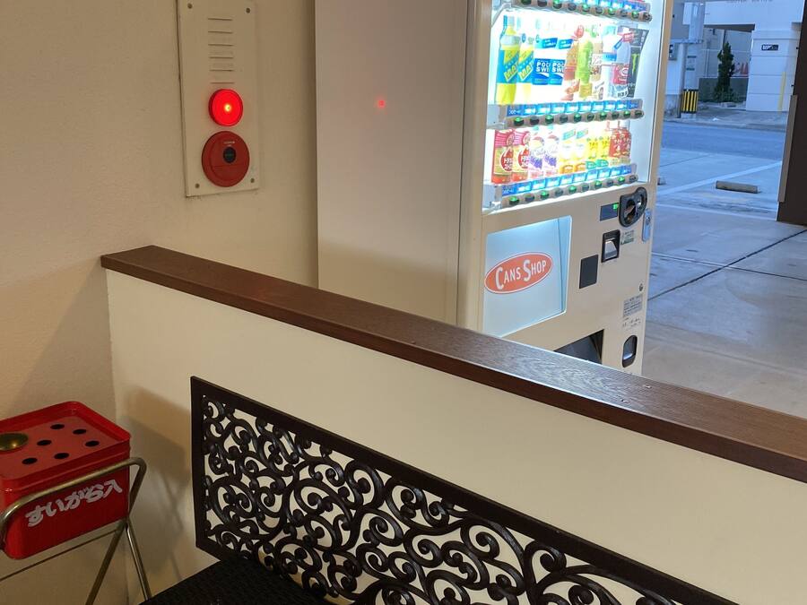 A vending machine with a variety of beverages and snacks, a red emergency button, and a metal chair with a decorative backrest.