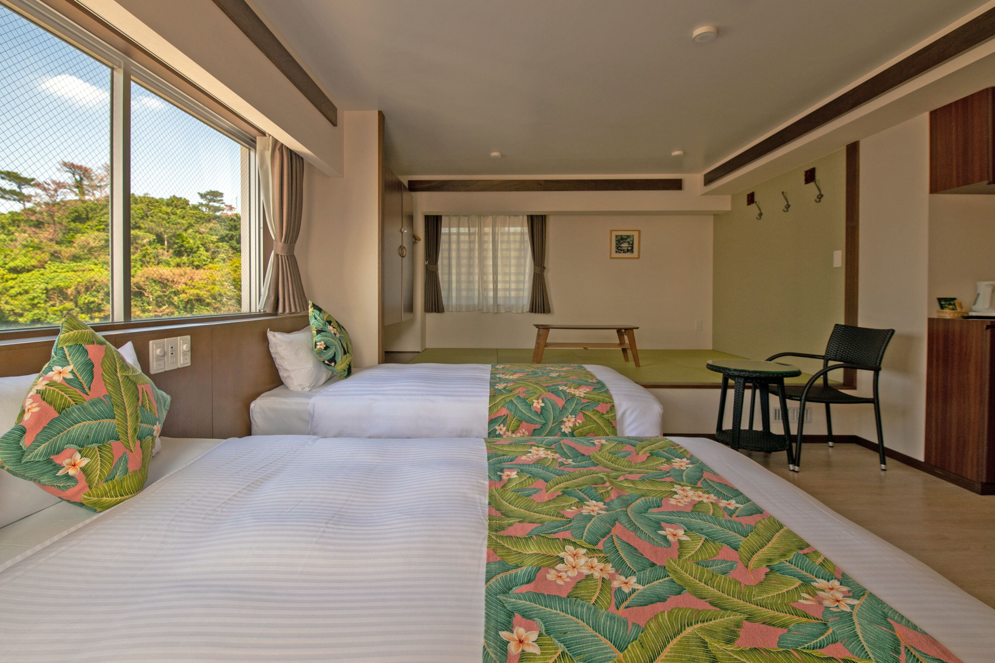 A hotel room with two beds, a small table, a chair, and a view of the outdoors.