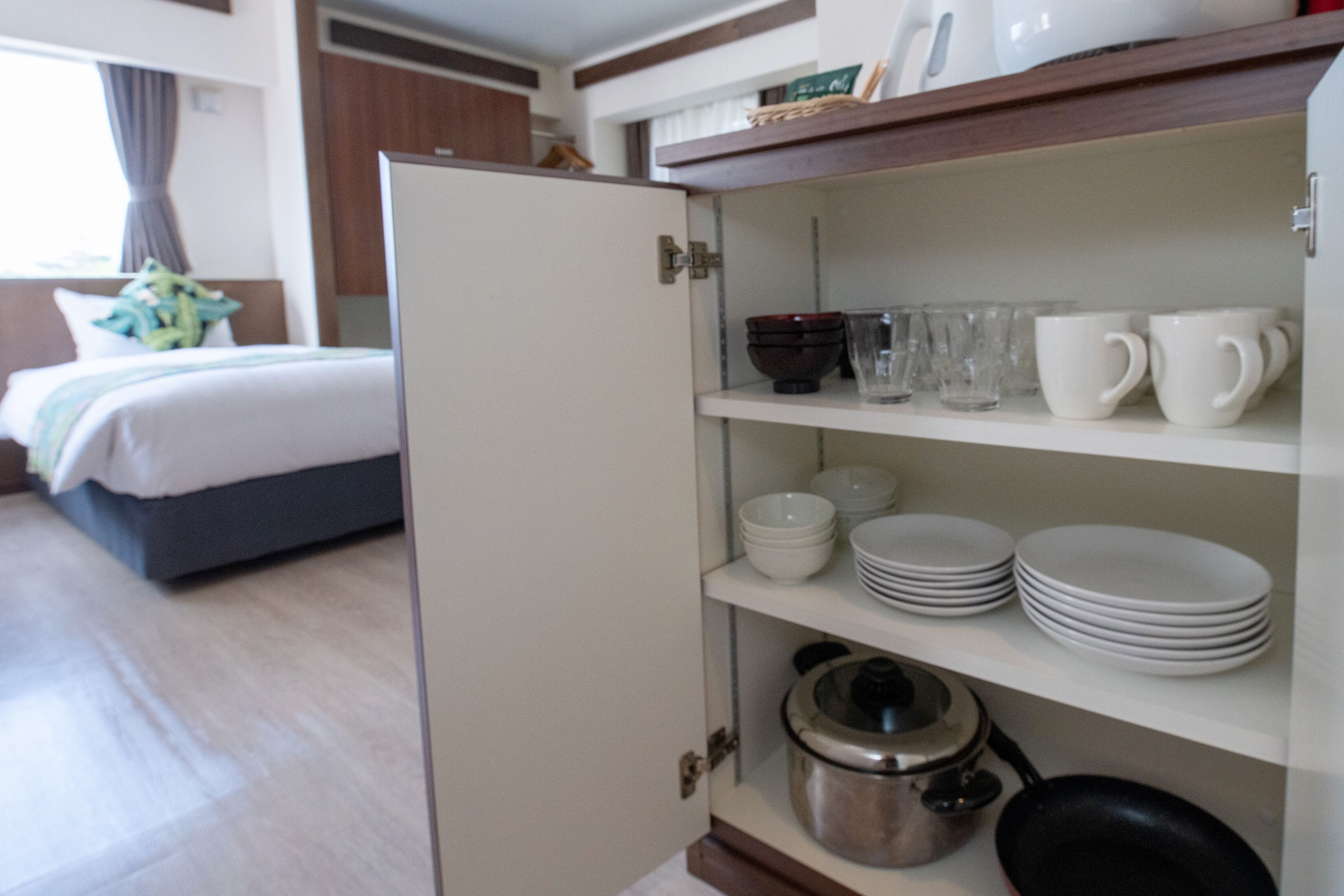 Una habitación de hotel con una cama, un armario blanco con vajilla y utensilios de cocina, y una ventana con cortinas.