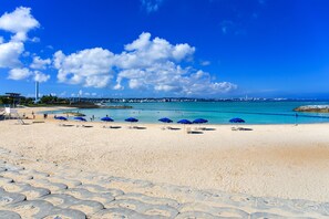 A beach with blue umbrellas, a clear sky, and a distant cityscape. - Aquaresort In Chatan (Chatan)