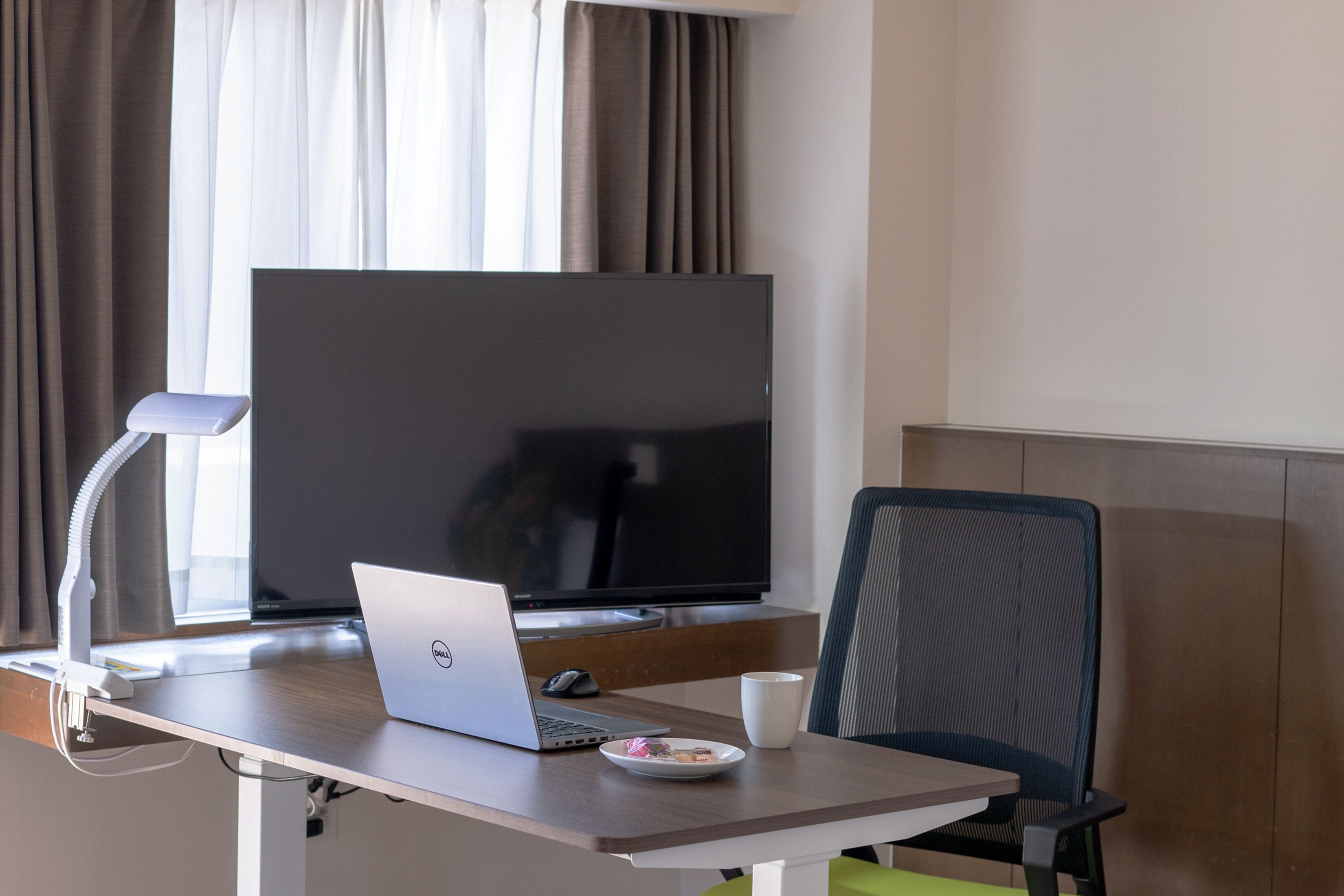 A hotel room with a desk, a laptop, a television, a chair, and a window with curtains.
