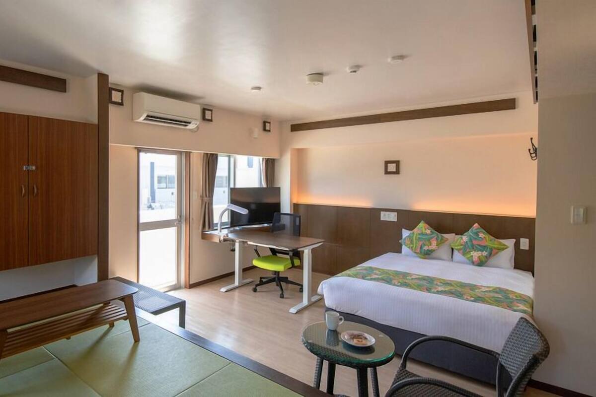 A hotel room with a bed, desk, chair, wardrobe, and a small table with a cup.