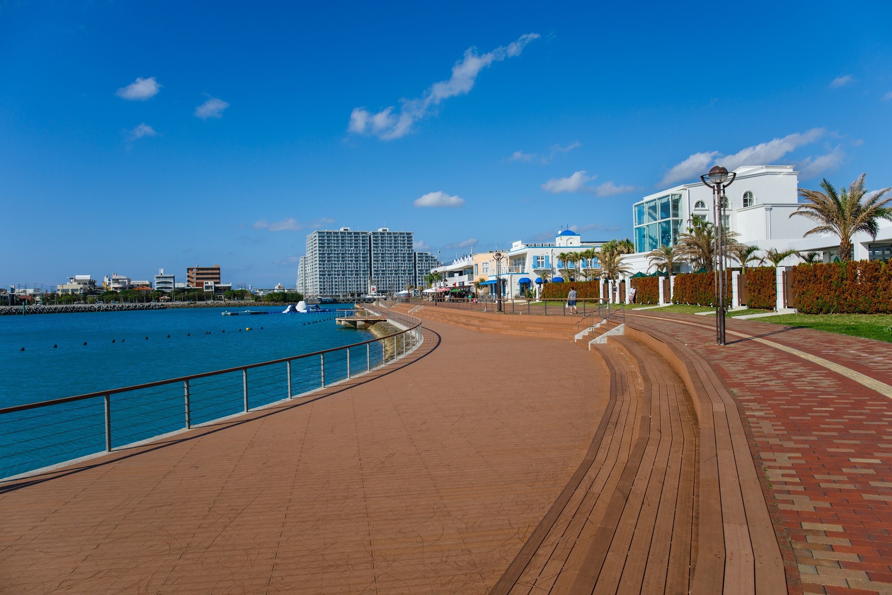 Un paseo marítimo con un camino de madera, cielo azul despejado y edificios modernos al fondo.