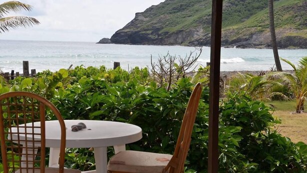 A covered outdoor seating area with a table and chairs, overlooking a coastal landscape with hills and the sea. - Hakamoui Plage (Ua Pou)