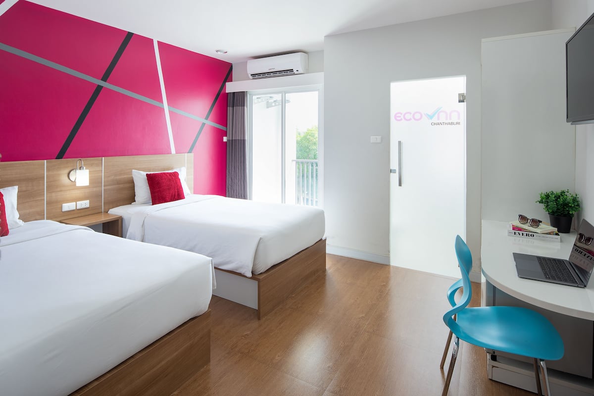 A hotel room with two beds, a desk with a laptop, a chair, and a view of the outdoors.