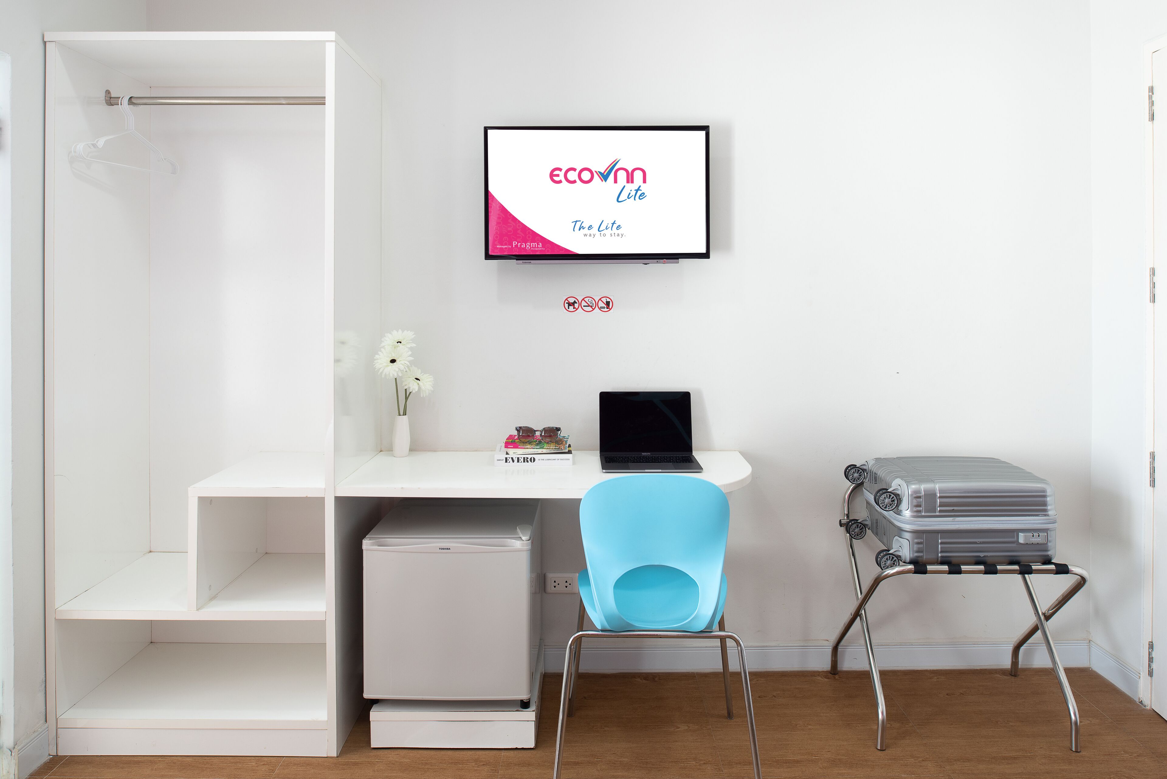 A room with a white desk, a laptop, a blue chair, a suitcase on a stand, and a shower area.