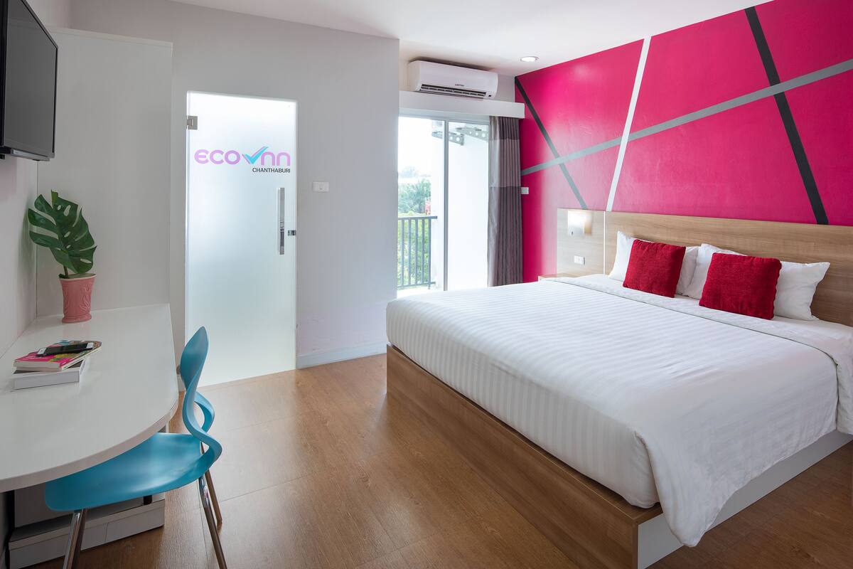 A modern hotel room with a large bed, a desk with a chair, a potted plant, and a view of the outdoors.