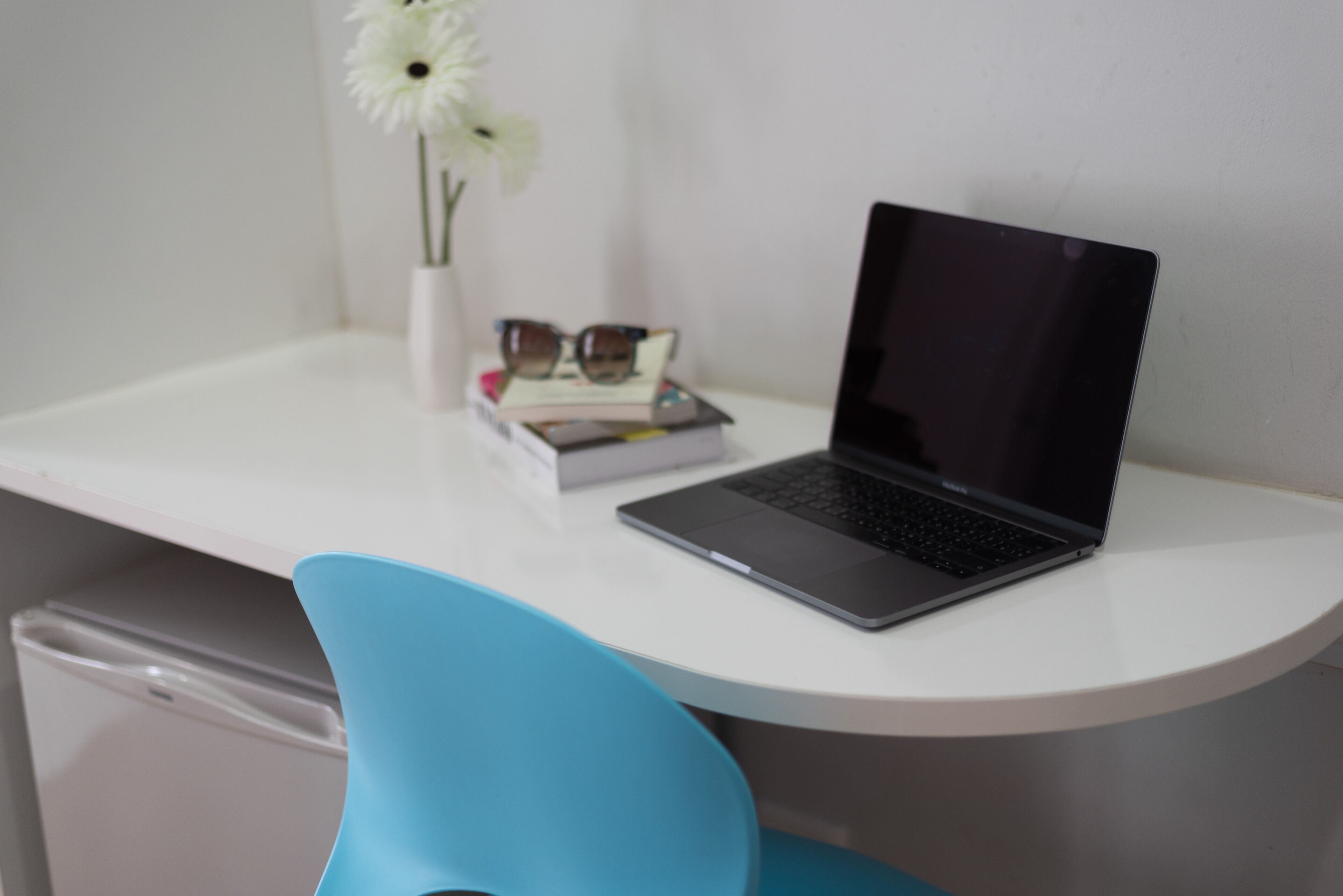 A laptop on a white desk with a blue chair, a white vase with a flower, and a book.