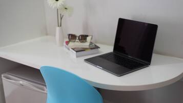 A laptop on a white desk with a blue chair, a white vase with a flower, and a book.