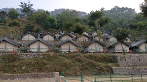 A row of simple, tent-like structures with a stone wall and green railings, set against a backdrop of trees and hills. - Camp Dogadda (Lansdowne)